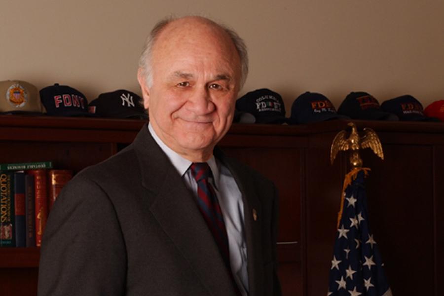 Former FDNY Commissioner Nicholas Scoppetta smiles as he poses for a portrait photo in a suit and tie. A row of FDNY baseball caps is visible on a bookshelf behind him.