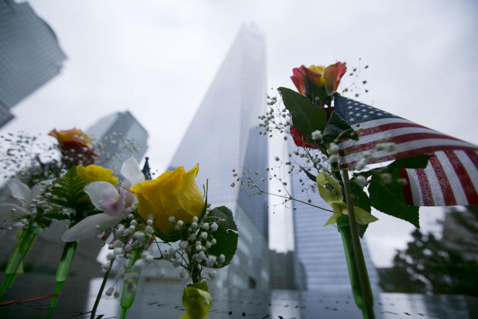 A small American flag and flowers, including yellow roses, stand at names on a bronze parapet at the 9/11 Memorial. One World Trade Center towers above on an overcast day.