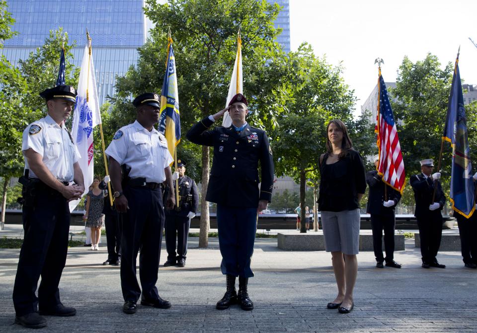 Medal of Honor recipient Ryan Pitts salutes as he stands in front of a color guard at Memorial plaza. Pitts is wearing a formal military outfit and is standing beside a woman and two uniformed police officers. Several trees on the plaza are behind him, as is One World Trade Center.