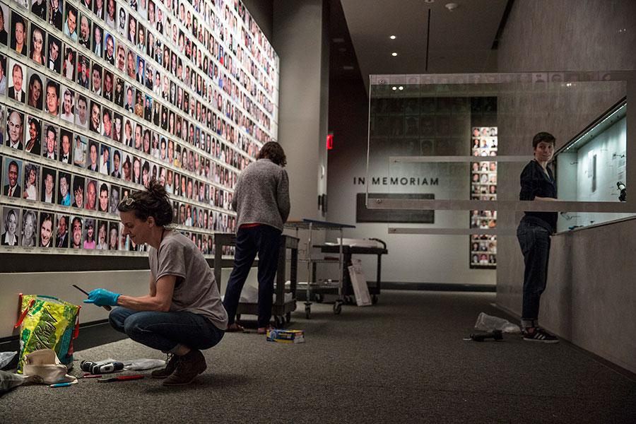 Three people install artifacts during off-hours at the 9/11 Memorial Museum. A wall featuring images of victims of the attacks towers over two women to the left.