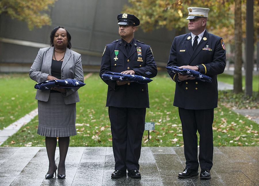Port Authority Chief Operating Officer Stephanie Dawson, FDNY Battalion Chief Joseph Duggan Jr., and NYPD Detective Nelson Vergara hold three folded American flags during a wreath-laying ceremony at the 9/11 Memorial.
