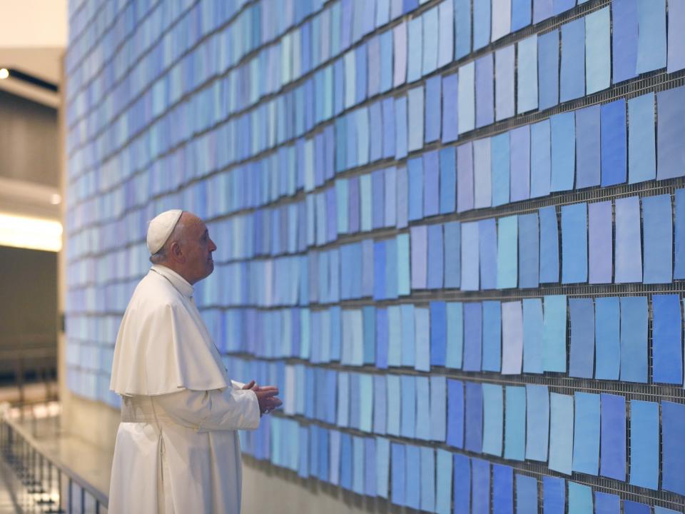 Pope Francis views “Trying to Remember the Color of the Sky on That September Morning” by artist Spencer Finch. The pope is wearing a white outfit and cap as he looks at the dozens of blue tiles that make up the artwork.