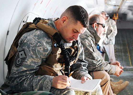Master Sgt. Bubba Beason wears a camouflaged outfit as he writes in a notebook while seated on a U.S. Air Force plane. Several other members of the Air Force sit beside him.