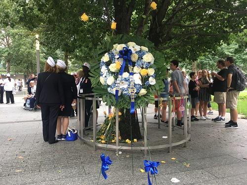 A white, yellow, and blue wreath laid by Blue Star Mothers stands next to the Survivor Tree at the Memorial.
