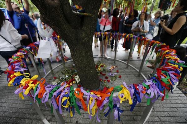 Dozens of ribbons in every cover of the rainbow are seen tied to a railing surrounding the Survivor Tree on the second anniversary of the Pulse nightclub shooting. Some in the background take photos of the tree as others tie ribbons on the branches of the tree.