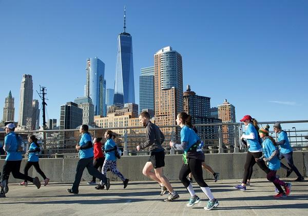 About a dozen runners are seen participating in the 9/11 Memorial & Museum 5K Run/Walk and Community Day. They are running on a concrete bridge with One World Trade Center and lower Manhattan behind them.