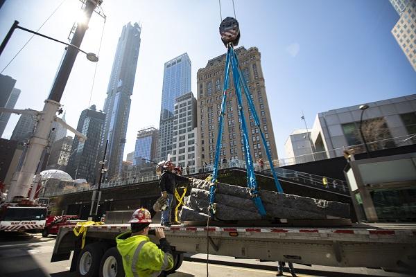 A 600-ton crane hoists a stone monolith to be placed at the 9/11 Memorial Glade on the Memorial plaza. Workers help position the monolith as it sits on the back of a flatbed truck.