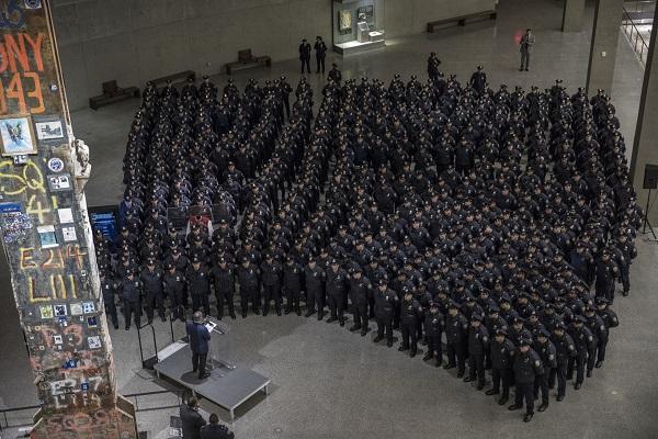Hundreds of NYPD recruits stand in Foundation Hall. A man is speaking to them from a stage beside the Last Column.