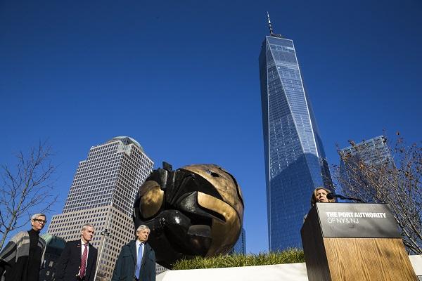 Alice M. Greenwald, the president of the 9/11 Memorial & Museum, speaks at a podium next to the Koenig Sphere in Liberty Park. One World Trade Center towers over her and others at the event on a cloudless day.