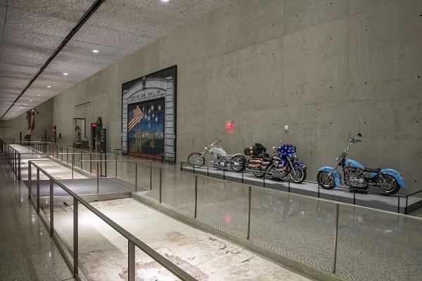 Three motorcycles and other Museum artifacts line the concrete wall of the Tribute Walk. The door of a firehouse painted with a patriotic mural is beside the motorcycles. The exposed box columns of the South Tower line the floor off to the left.