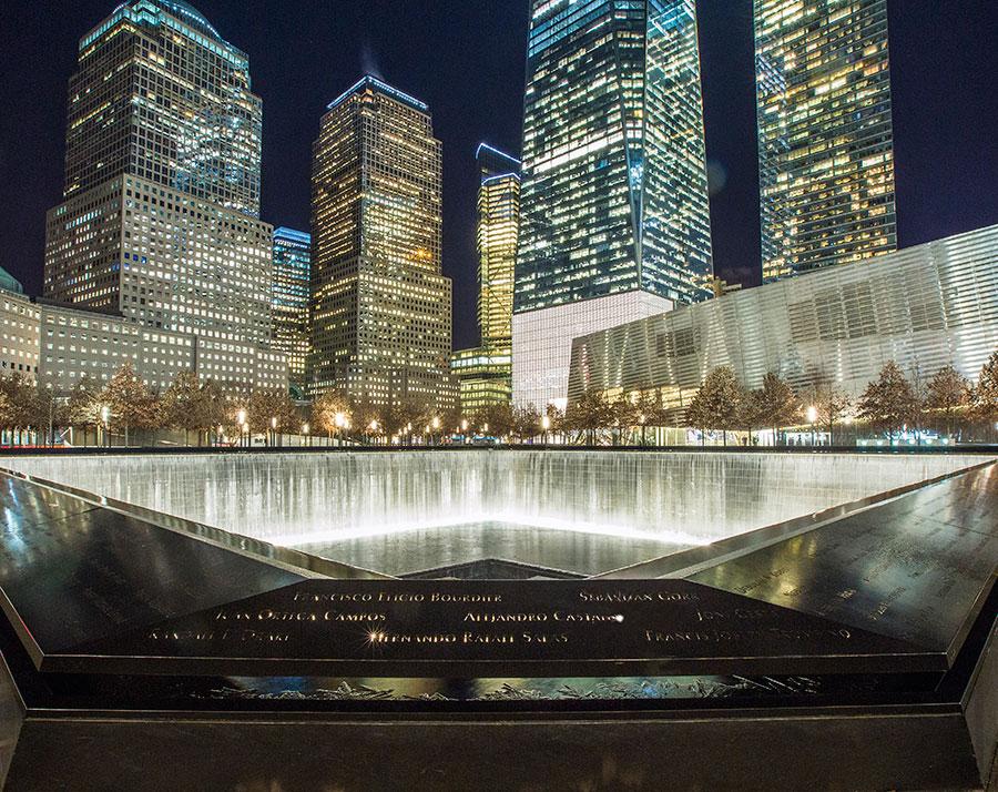 A reflecting pool of the 9/11 Memorial is seen illuminated at night. Surrounding buildings, including One World Trade Center, are lit up in the background.