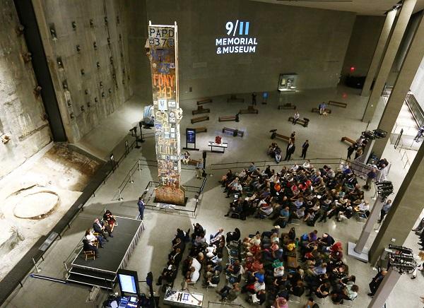 In this view over Foundation Hall at the 9/11 Memorial Museum, a sitting audience watches four people onstage beside the Last Column. The massive space includes the slurry wall and an area with about a dozen benches.