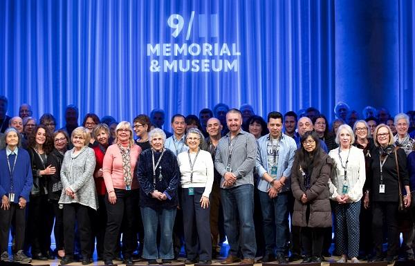 Dozens of volunteers, primarily older adults, are seen onstage at the Museum auditorium during the National Volunteer Week ceremony and reception. A blue curtain behind them has the 9/11 Memorial & Museum logo projected on it. 