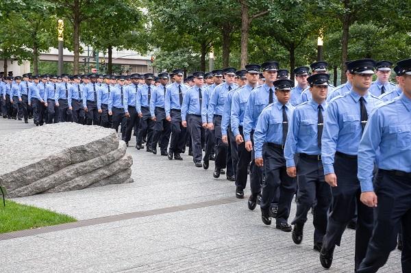 Dozens of FDNY probationary firefighters in formal, blue uniforms pass through the 9/11 Memorial Glade.