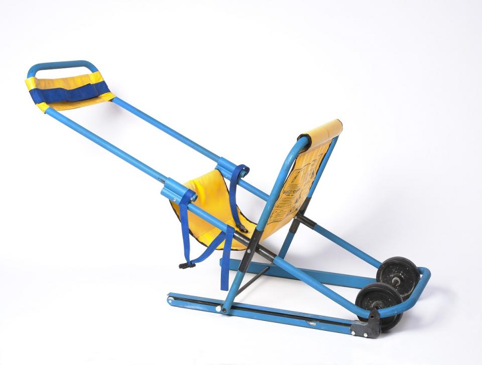 A blue and yellow evacuation chair used on 9/11 is displayed on a white surface in the Museum. The chair was used by to evacuate a quadrplegic man, John Abruzzo, from the North Tower. The chair includes wheels and sled-like components that make it easier to descend stairs.