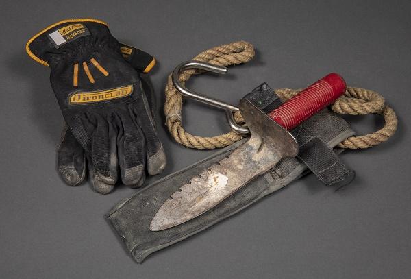 Tools and a pair of gloves belonging to George Torres of FDNY Squad 41 are displayed on a gray surface. The tools include a garden trowel with a red handle that Torres used to dig at Ground Zero.