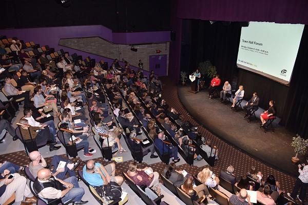 A town hall meeting of onePULSE Foundation is held at an auditorium. Dozens of audience members sit as they watch six people onstage.