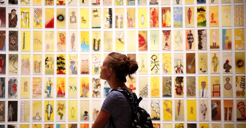 A woman with a backpack walks through the exhibition “Rendering the Unthinkable: Artists Respond to 9/11.” Dozens of small artworks are displayed on the wall in front of her.