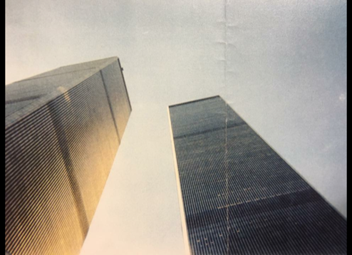 A photograph by Jeffrey Keating shows the Twin Towers from below on September 10, 2001. Sunlight shines on one of the towers. The other is cloaked in shadow.