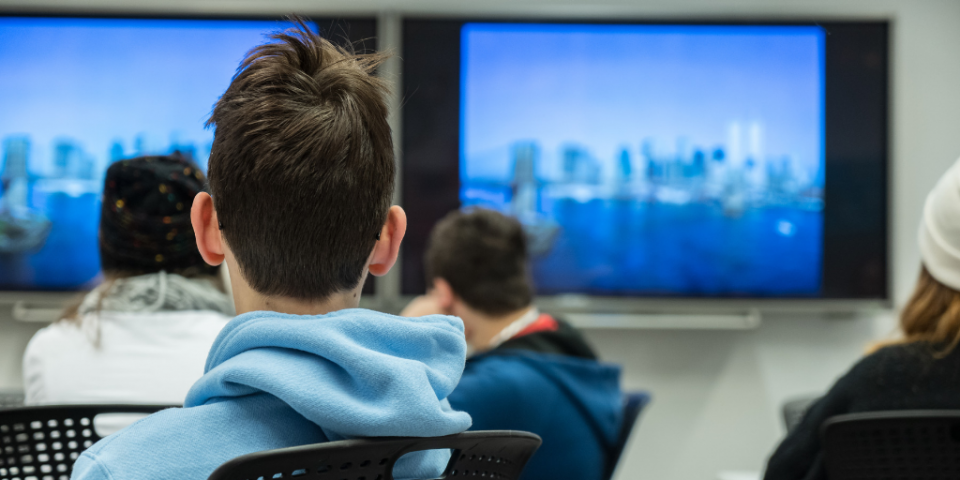 A student in a hoodie sit with his back to the camera in the Museums’ Education Center. He and other students are watching television screens that are showing lower Manhattan.