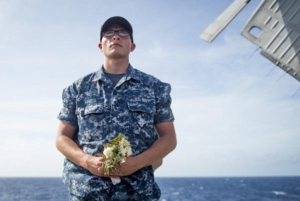 A man in blue U.S. Navy camouflage stands on a ship while holding a posse of flowers. An expansive view of water and sky is behind him.