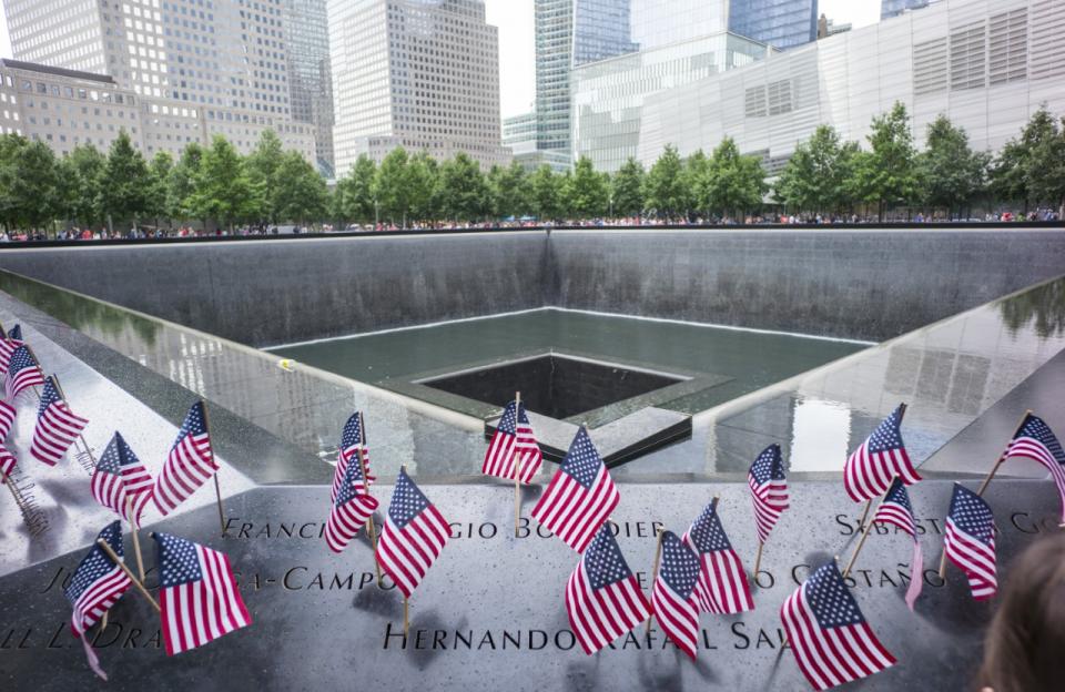 American flags are placed in the names of the 9/11 Memorial parapets. The center of the Memorial pool is visible behind them.