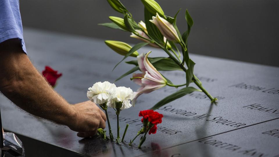 A family member places a white carnation in a name on a memorial panel.  His arm is extending into the frame from the left side as he add a carnation to row flowers placed inside the name..    