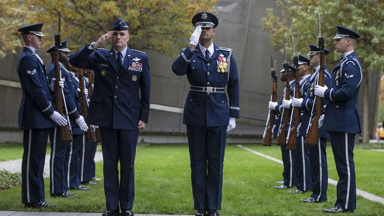 Members of the U.S. Air Force drill team outfitted in navy blue dress uniforms stand in two identical rows with rifles, while two members stand side-by-side in the foreground saluting away from the other members. In the background is the green lawn of the 9/11 Memorial plaza and trees that have begun to change color for the fall.