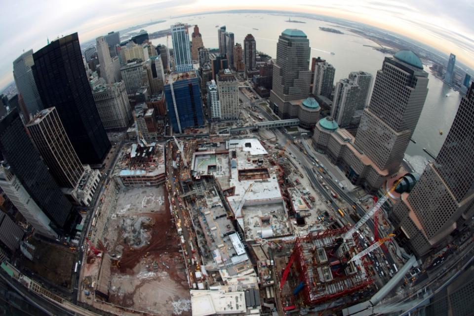 An aerial view shows construction at the 9/11 Memorial site. The buildings of lower Manhattan and New York Harbor are visible in the distance.