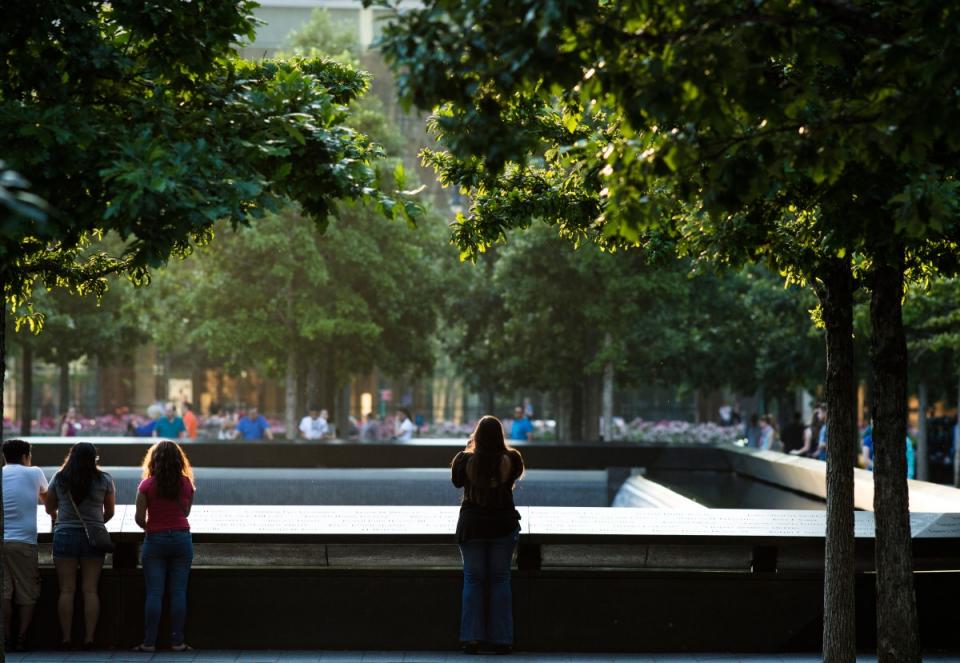 Four visitors stand beside the reflecting pool on a warm evening. The trees of Memorial plaza line the reflecting pool around them.