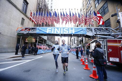 Participants cross the finish line at the 2015 9/11 Memorial 5K Run and Walk. The participants run under a sign saying “Finish” and adorned with more than a dozen American flags. 