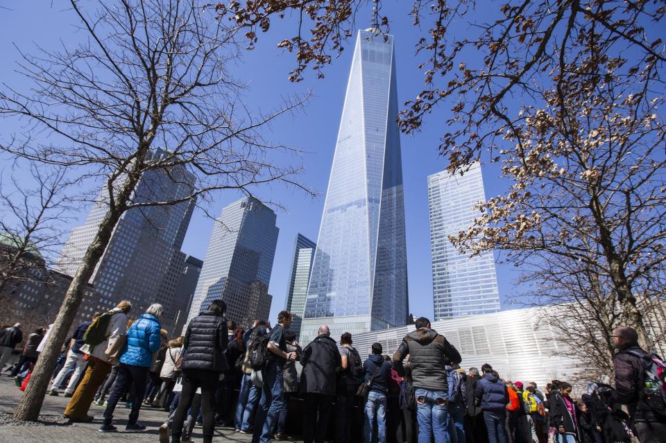 One World Trade Center towers over visitors at the 9/11 Memorial plaza on a sunny day. Swamp white oak trees stand over the visitors in the foreground.