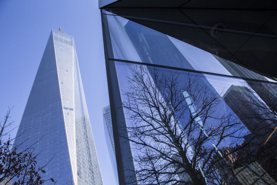 One World Trade Center rises over the 9/11 Memorial plaza. The glass facade of a building reflects a leafless tree in the foreground.