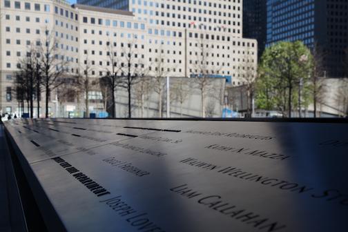 Shadows fall over names inscribed on bronze parapets at the 9/11 Memorial. World Financial Center buildings are in the background.