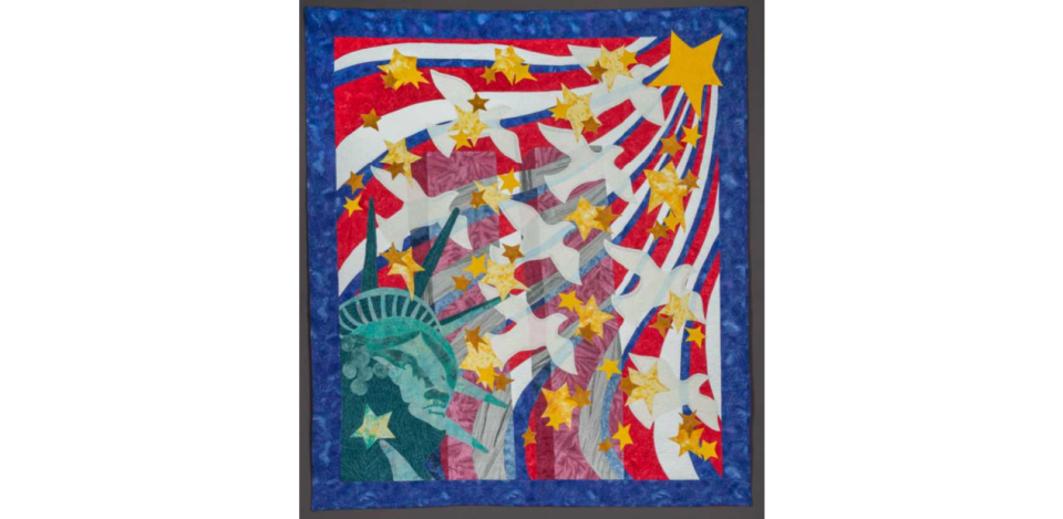 Handmade textile quilt depicts the Twin Towers at center, ghostlike in their transparency. The crowned head of the Statue of Liberty is represented in the lower left corner of the quilt, and from it white doves and yellow stars emanate. These images are set against waves of red, white, and blue. 
