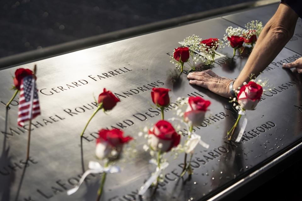 A man braces an arm on the names parapet, alongside red flower tributes.