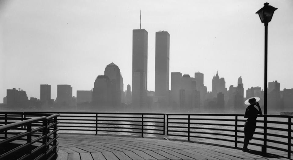 A woman in silhouette leans against a lamppost on a curved boardwalk, facing the view of lower Manhattan in the background. The Twin Towers, at the center of the skyline, rise into the cloudy sky.