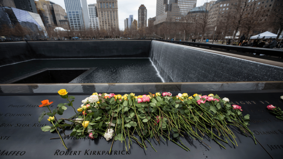 Dozens of roses are laid upon the North Pool names panel of the portion of the 9/11 Memorial dedicated to the victims of the 1993 World Trade Center attack.  A few roses are standing up after being placed inside the names of 1993 victims as water cascades down the side of the Memorial on an overcast day.