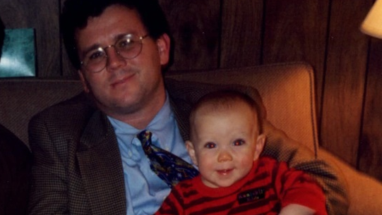 A young man, wearing glasses and a jacket and tie, holds his rosy-cheeked baby daughter on his lap as they both smile sweetly for the camera.  The family resemblance is apparent in their upturned smiles and the baby's smiling eyes convey her love and delight to be in the company of her dad.  