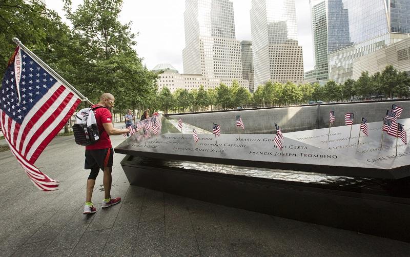 A man in black shorts and a red top carries a large American flag as he places a smaller one alongside names on the 9/11 Memorial.
