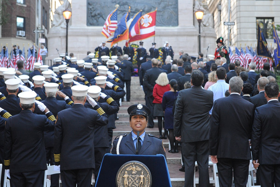 A woman in a navy blue firefighter's uniform speaks at a podium. Behind her,  the backs of program attendees saluting six uniformed men on a stage.