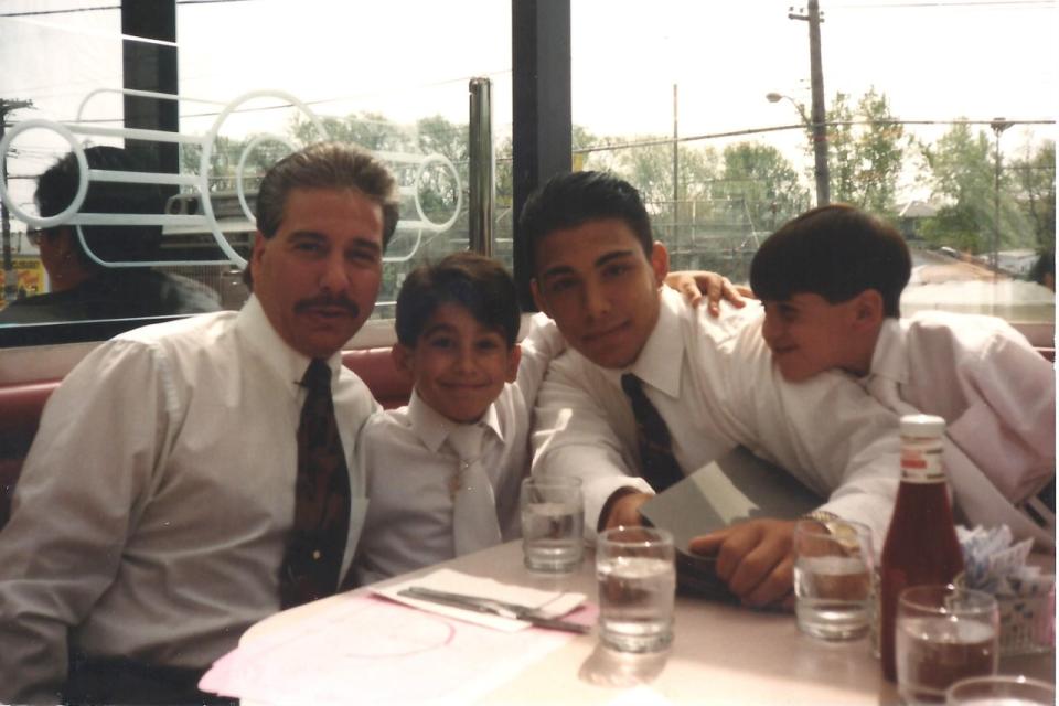 An adult male in a white shirt and black tie, with dark hair and a mustache, smiles next to three younger boys, also in white shirts with black ties, at a diner table.