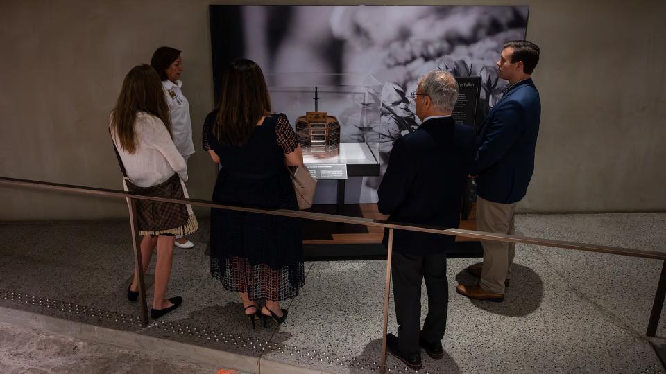 Five formally-dressed adults stand, backs to camera, looking at a wooden octagonal memorial stand covered with engraved black and brass plaques.